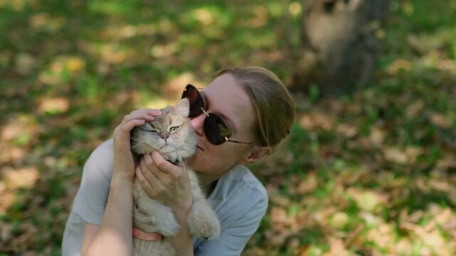 Woman holds cat gently while sitting under trees shade. She hugs her pet tenderly. Cat looks calm, eyes half-closed in trust. Sunlight filters through leaves, soft and warm.