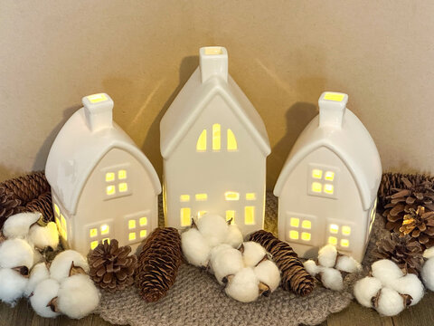 Glowing white ceramic village houses styled with cotton bolls and pinecones in a neutral palette, creating a cozy Scandinavian display for autumn, winter, Thanksgiving and Christmas.
