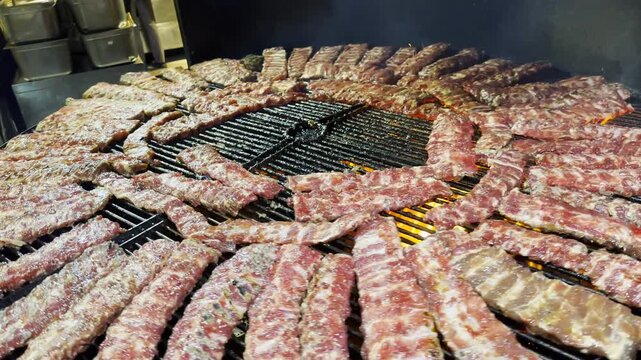 Pork ribs rotate on a metal grill slowly roasting at restaurant kitchen. The meat sizzles over open heat developing rich golden crust. The focus on cooking process, texture and appetizing appearance