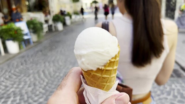 POV of male hand holding an ice cream cone while walking along city street. Blurred pedestrians pass by in the background, creating a lively urban atmosphere and emphasizing the treat in arm