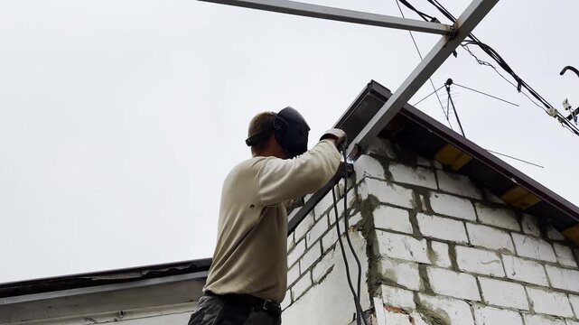 Manual laborer in protective mask welds iron detail at construction. Professional young repairman with welding machine working at the site. Male welder operating with metal on house. Close up