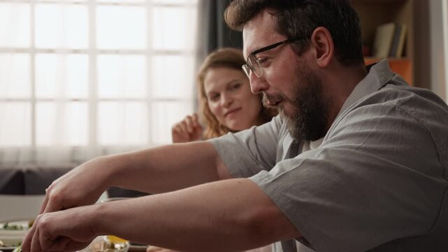 Caucasian middle aged man cutting roasted meat during family dinner with Caucasian woman smiling in background. Perfect for holiday celebration promos or food brand commercials