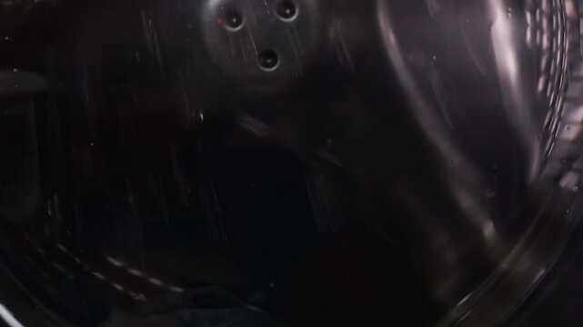The washing machine is washing clothes, the drum with water and detergent is rotating. Medium shot.