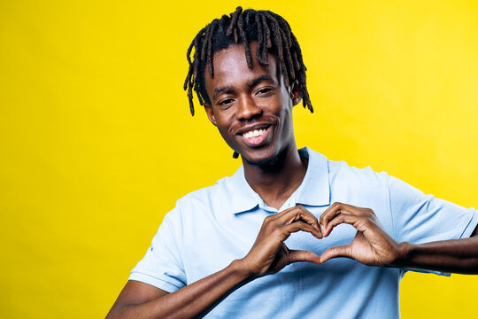 Young black man with dreadlocks smiling brightly, forming a heart shape with his hands on a vibrant yellow background
