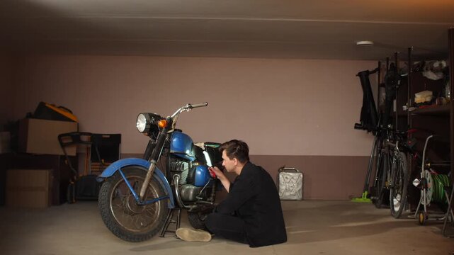 Man feeling headache and frustration while attempting to fix a broken, non functioning old blue motorcycle parked in a cluttered home garage