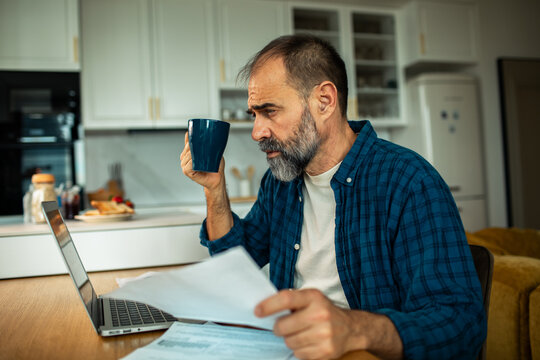 Worried middle-aged man reviewing bills at laptop in home kitchen