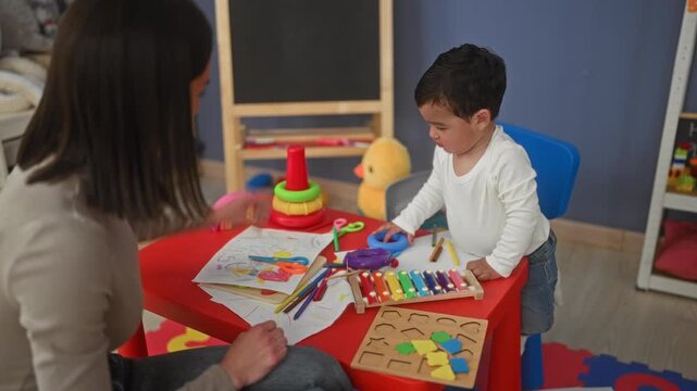 Teacher guides little boy playing with xylophone and stacking rings, boy reaches for a ring on a red table inside a childcare building; curiosity bonding.