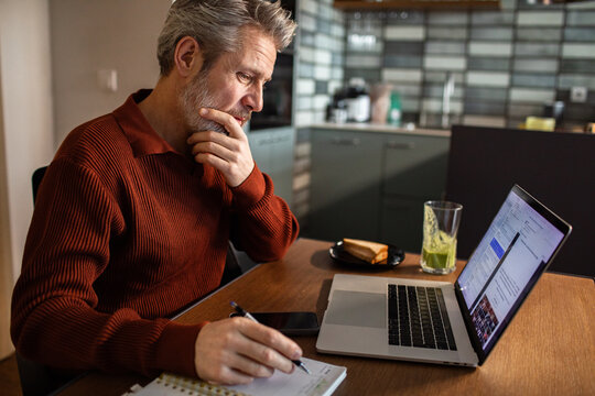 Middle-aged man taking notes while working on laptop at home kitchen table