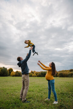 Photo portrait of dad throwing little boy up in the air while pregnant mom watching outdoors in autumn field. Family having fun together during a walk in nature under cloudy sky.