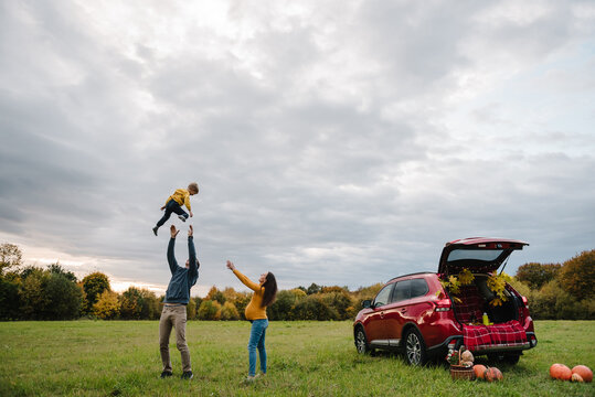 Photo portrait of dad throwing little boy up in sky near car trunk decorated with autumn leaves, pumpkins, blanket, basket on grass. Pregnant mom watching outdoors in autumn field during road trip.