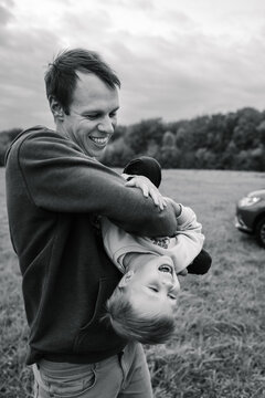 Black and white photo portrait of dad holding laughing little boy upside down outdoors in autumn field. Father and son having fun together during a walk in nature.