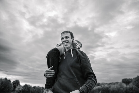 Black and white photo portrait of smiling dad carrying little boy on his back outdoors in autumn field. Father and son having fun together during a walk in nature under cloudy sky.