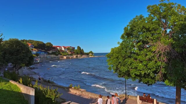 Scenic coastal view of the Nessebar Old Town on the Black Sea, Bulgaria. This 4K (UHD) video captures a tranquil seaside promenade with gentle waves, lush green trees, and traditional red-roof houses 