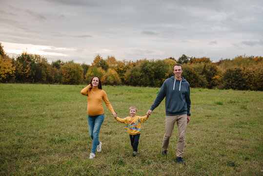 Family walking together holding hands outdoors in autumn field. Pregnant mom, dad, and little son having a walk in nature.