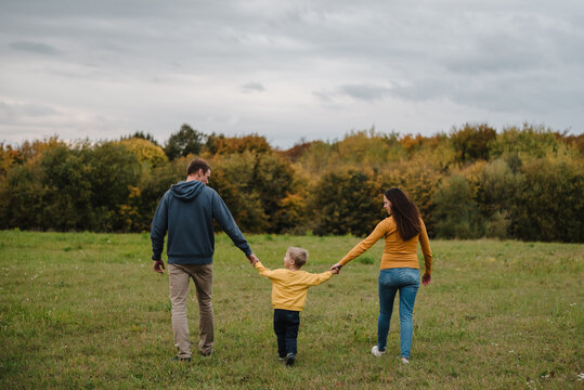 Family walking together holding hands outdoors in autumn field, back view. Dad, mom, and little son having a walk in nature.