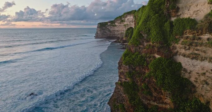 Tropical Bali coastline aerial view, dramatic limestone cliffs draped in lush green vegetation meeting turquoise Indian Ocean with gentle waves and pastel sunset sky. Drone flight evening landscape