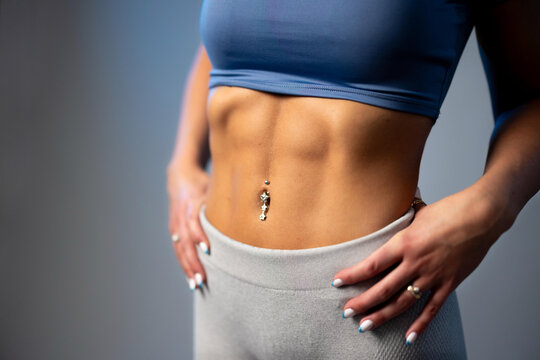 Fit woman showing defined abs in blue sports top and gray leggings with belly button piercing