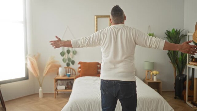 Man standing on bed with arms outstretched and open palms in a sunlit building; contentment morning routine.