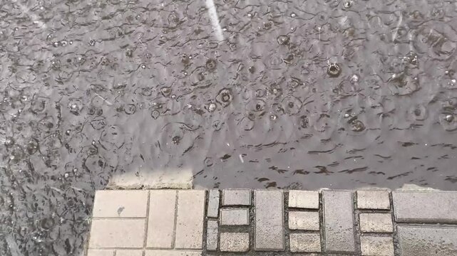 Raindrops falling on wet asphalt creating ripples and splashes beside a paved sidewalk. Atmospheric rainy weather background with water texture, urban street surface, and storm mood.