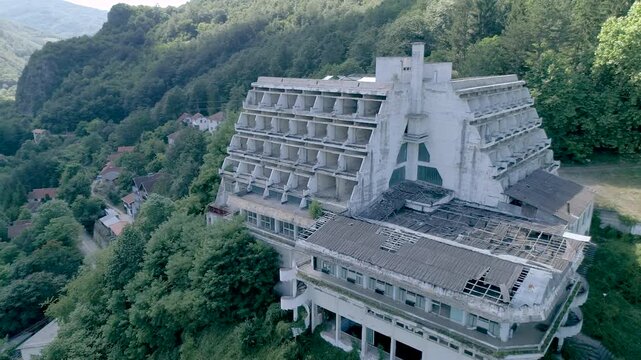 Close aerial drone view of a large abandoned concrete hotel in a forested mountain valley, showing damaged roof, empty windows and deserted resort architecture.