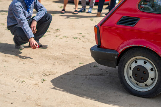 Man crouching near a red classic Fiat 126p car measuring exhaust noise level in decibels using a sound level meter on sandy ground in bright natural daylight sunlight.