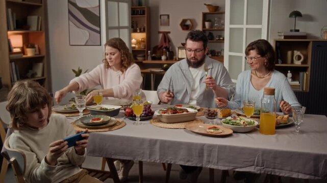 Caucasian family gathering around dinner table while man cutting roasted meat and kid playing on smartphone. Perfect for family lifestyle commercials or technology distraction concepts