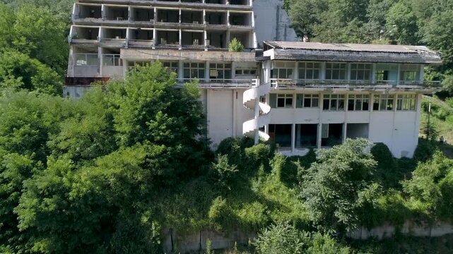 Aerial drone shot rising past an abandoned multi storey hotel building, revealing dense green forest and mountain landscape around the deserted resort.