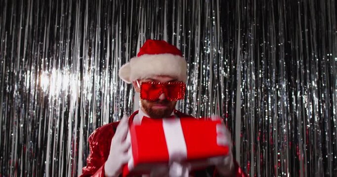 Christmas funny dance of Santa dancer with red gift box in front of tinsel curtains. Happy man with festive sequin costume, glasses and Santa hat dancing and spinning, receiving present on party