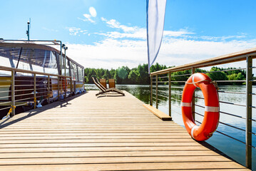 A pontoon boat is moored to a wooden dock for relaxation by the tranquil waters surrounded by green trees. © Natallia