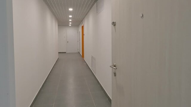 First person perspective of an apartment exit. Scene begins in a white walled hallway with an intercom and tiled floor. Camera pans left through an open door into a long narrow building corridor