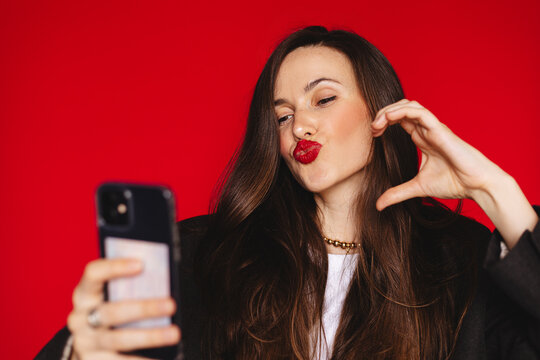 Close-up portrait of attractive young woman with long dark hair and red lipstick making a kissy face while looking at smartphone screen and showing half heart hand sign isolated red studio background
