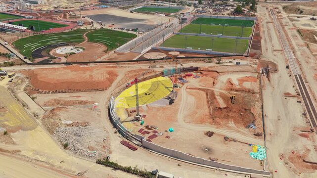 Expansive aerial view showcasing the active construction of a modern sports complex, with workers and machinery developing an amphitheater and surrounding athletic fields in a large-scale project
