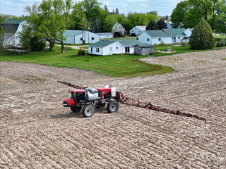 Red self-propelled field sprayer applying herbicide to a farm field with a farm scene in the background. © dvande