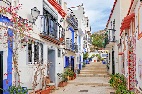 Narrow cozy street with steps, white houses, potted plants in historic Santa Cruz neighborhood of Alicante Old Town, located near Santa Barbara Castle