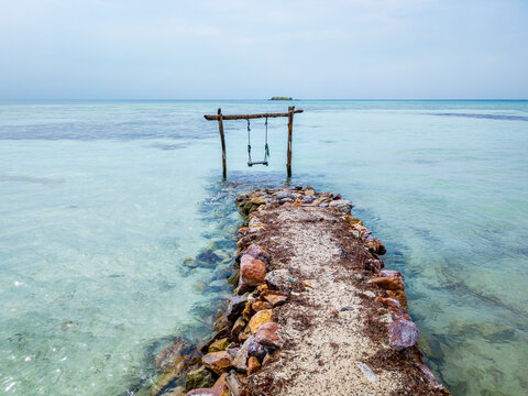 Rustic swing structure in the shallow, turquoise ocean water beside a sandy path, creating a serene and idyllic scene for relaxation at Bobby Beach in Karimunjawa, Indonesia