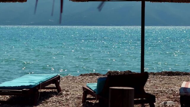 Empty Sunbeds Under Umbrella Canopy On Pebble Beach Overlooking Sparkling Turquoise Sea Water. Summer Resort Vacation 