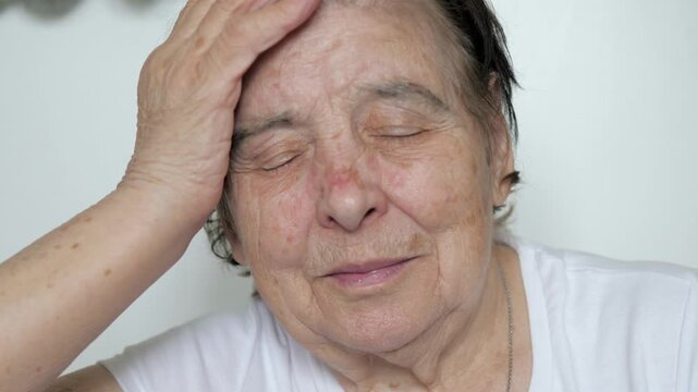 Upset elderly woman 80 or 90 years with deep wrinkles holding head and closing eyes, experiencing painful headache or migraine. Old female feeling stressed and tired while isolated on white background