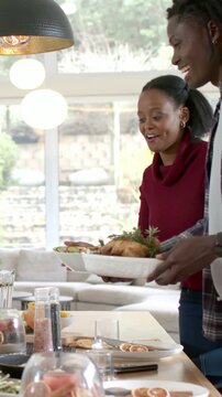 Vertical video: Entering right, couple in burgundy, plaid placing roast on dining table for serving