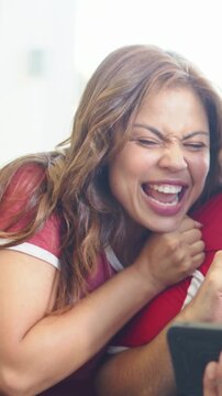 Vertical video: Leaning woman in red tee cheering on couch after goal on tablet, with soccer ball