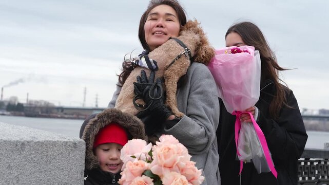 A woman holding a curly poodle, a teenage girl holding a pink bouquet of flowers and her young son on a calm beach, under a soft overcast sky with a view of the coast in the distance, a casual