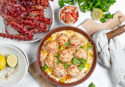 CRABMEATBALLS crab meatballs in white creamy sauce in red pan, whole king crab, cilantro, lemon and white wine on white background