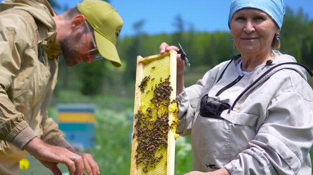A cinematic medium shot of a happy senior female beekeeper smiling at the camera while holding a fresh honeycomb frame at an apiary.
