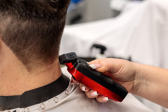 Hairdresser using a red electric hair clipper to trim a man hair in a professional barbershop, close up view of the professional hair cutting process today at the salon