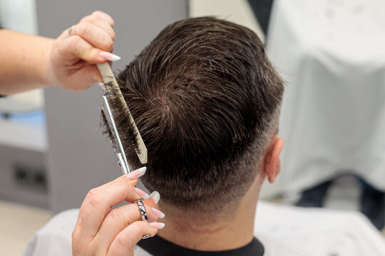 Barber using scissors and a comb to cut a man hair at the back of the head, professional hair cutting technique performed in a busy barbershop hair salon today afternoon