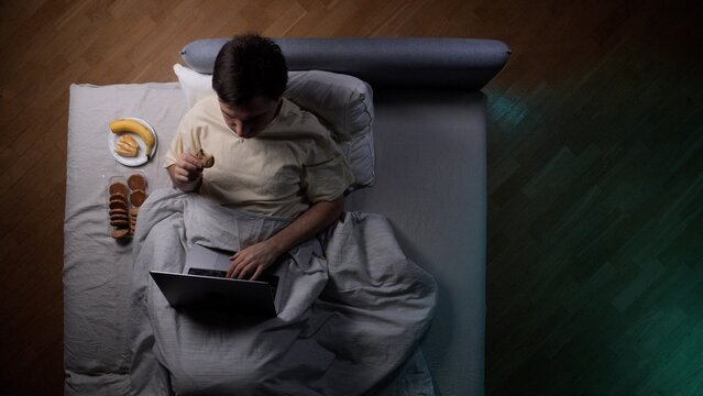 Young man lying in bed, using a laptop, and eating cookies and a banana, portraying a concept of late night work, unhealthy lifestyle, or digital addiction