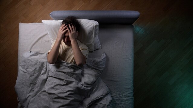 Young man lying in bed late at night, covering his face with hands, experiencing headache, feeling restless and frustrated due to inability to sleep