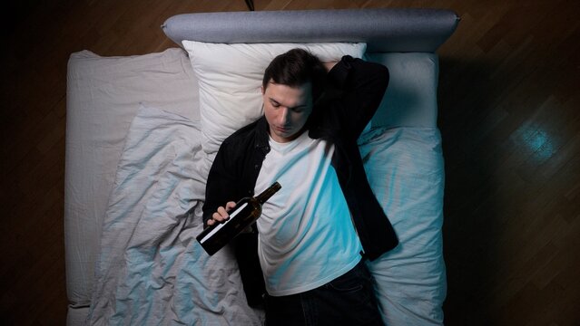 Young man lying on his back in bed, dressed in a t shirt and shirt, holding an empty bottle in his hand, experiencing feelings of loneliness and despair