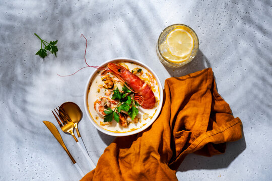 Creamy seafood chowder soup with prawn, mussels, cutlery, napkin, glass on concrete background. Summer seafood restaurant layout background, elegant gourmet dining concept.
