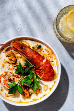 Seafood chowder soup with jumbo prawn, mussels in bowl with lemon drink on surface. Vertical layout gourmet seafood banner background, Mediterranean cuisine dish concept.