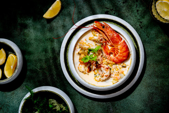 Seafood chowder soup with jumbo prawn, mussels, and lemon slice in hard light. Elegant summer lunch concept, on dark green background with refreshing drink.
